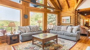 A log cabin interior with a large window, a sofa, a coffee table, and a rug. - Deep Creek Escape | Fireplace + Hot Tub (McHenry)