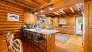 A log cabin interior with a kitchen featuring wooden cabinets, a central island with bar stools, and a dining area with wooden chairs. - Deep Creek Escape | Fireplace + Hot Tub (McHenry)