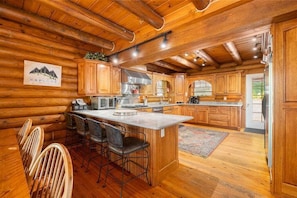 A log cabin interior with a kitchen featuring wooden cabinets, a central island with bar stools, and a dining area with wooden chairs.