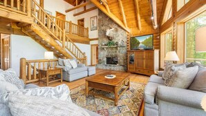 A log cabin interior with a stone fireplace, a wooden staircase, and a television. - Deep Creek Escape | Fireplace + Hot Tub (McHenry)