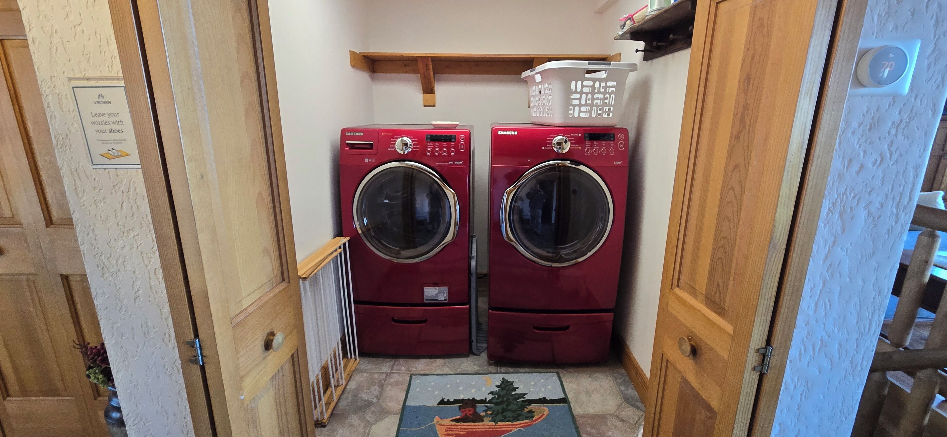 Una lavandería con dos lavadoras Samsung rojas, un estante de madera con cestas de ropa y una alfombra pequeña con un diseño de árbol de Navidad.