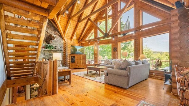 A log cabin interior with a wooden staircase, a fireplace, a sofa, and large windows. - Deep Creek Escape | Fireplace + Hot Tub (McHenry)