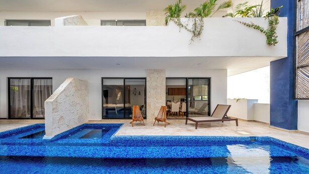 Un área de piscina exterior moderna con una estructura de piedra, sillas de descanso y un comedor con paredes de vidrio.