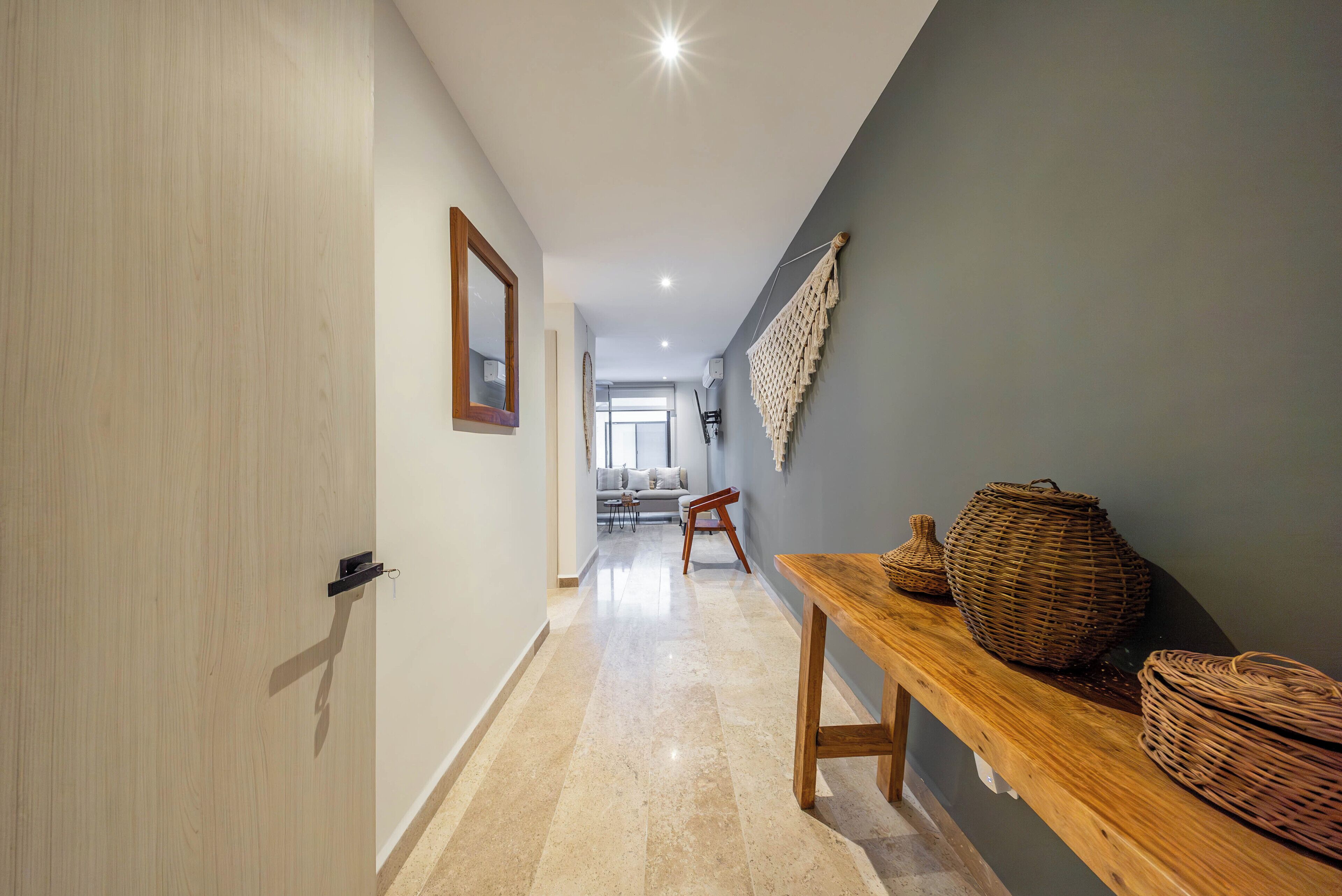 Un pasillo con puerta de madera, un espejo, cestas de mimbre sobre una mesa y vistas a una sala de estar con sofá y sillones.