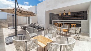 Un patio al aire libre con sillas de mimbre, una mesa de madera y un paraguas grande. También hay sillones reclinables y una zona de bar con taburetes.