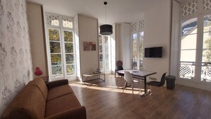 A spacious living room with a brown sofa, a dining table with chairs, a TV mounted on the wall, and large windows with decorative glass panels.