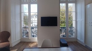 A room with a large window, a flat-screen TV mounted on the wall, a wooden floor, and a chair.