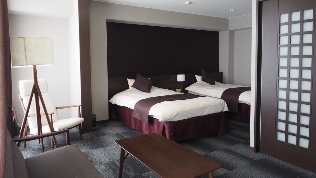 A hotel room with two beds, a desk, a chair, and a lamp.