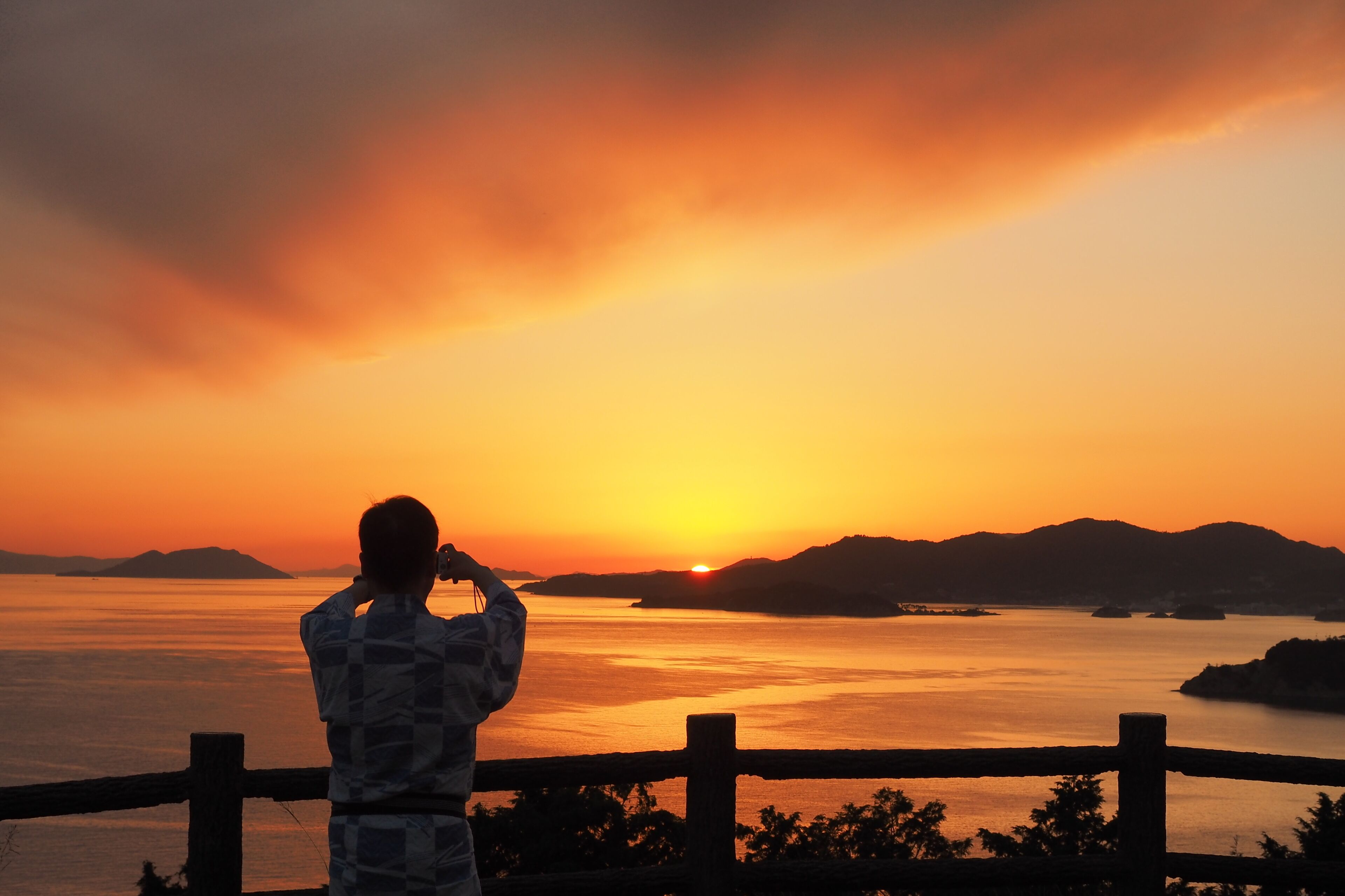 A person taking a photo of a sunset over a body of water with mountains in the background.