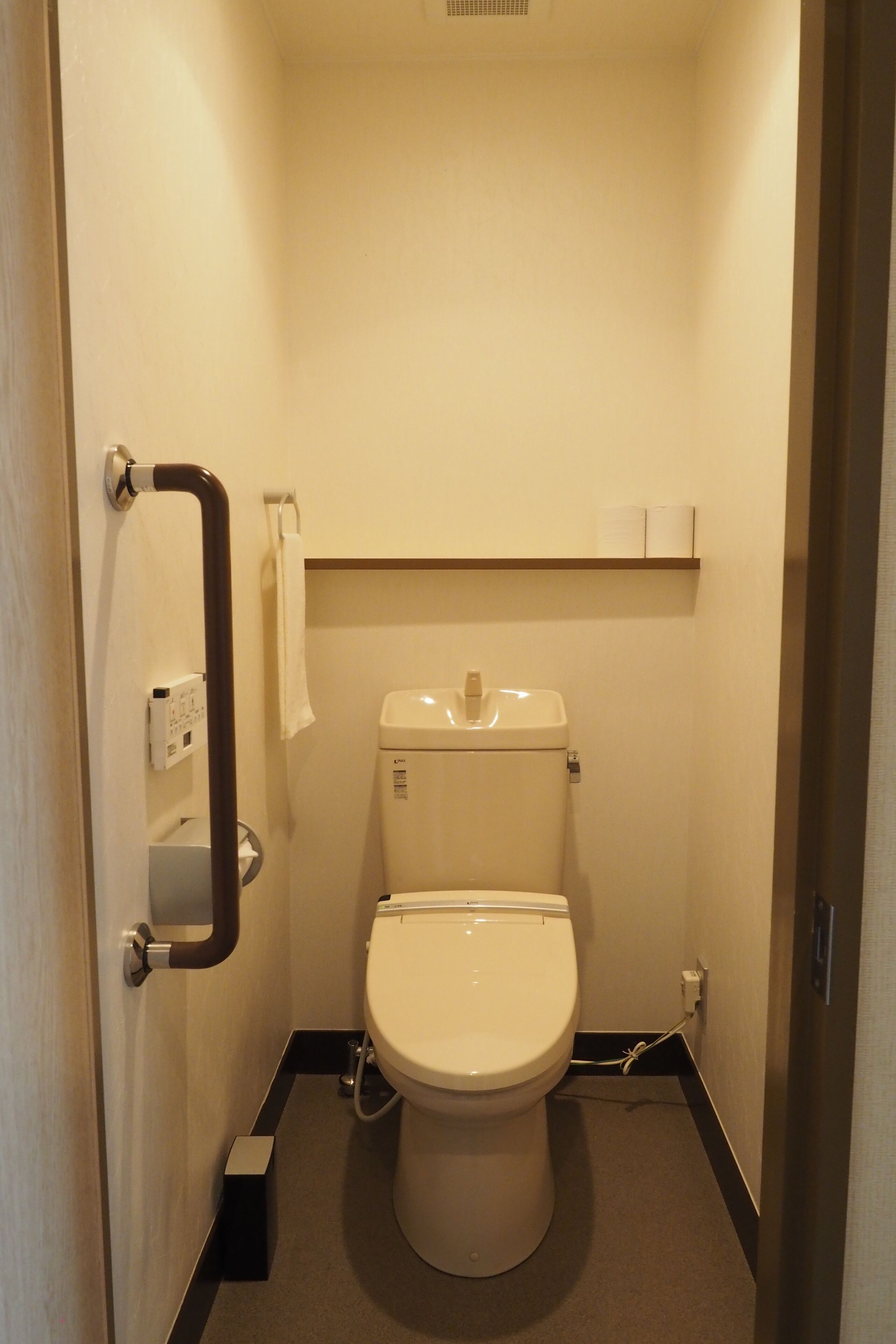 A small, compact bathroom with a toilet, a handrail, and a shelf above the toilet.