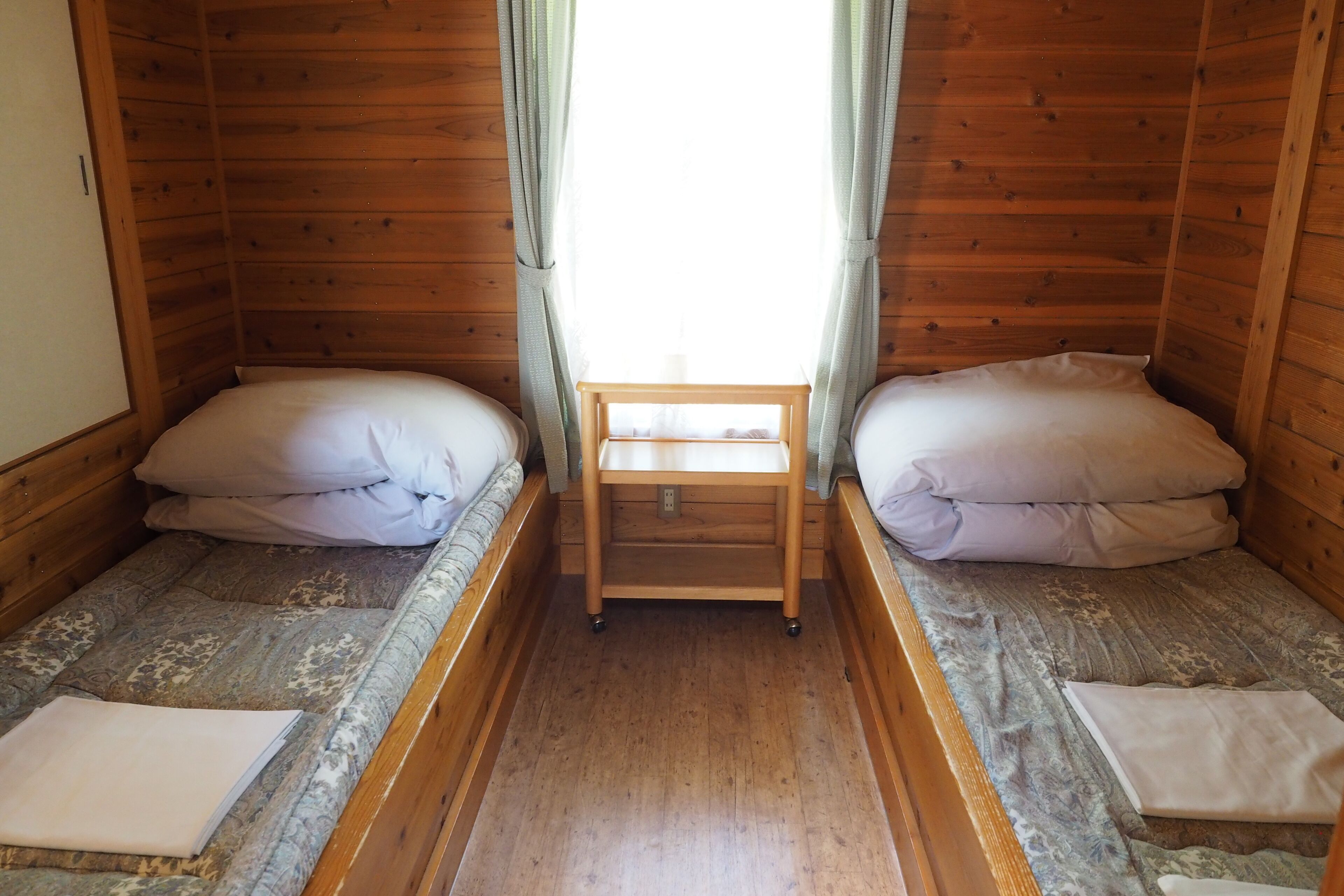 Une chambre avec deux lits simples, une petite table en bois et une fenêtre avec des rideaux.
