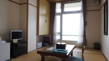 Una habitación japonesa tradicional con suelo de tatami, una mesa baja con cojín, un televisor sobre un mueble y una puerta corrediza con cortinas.