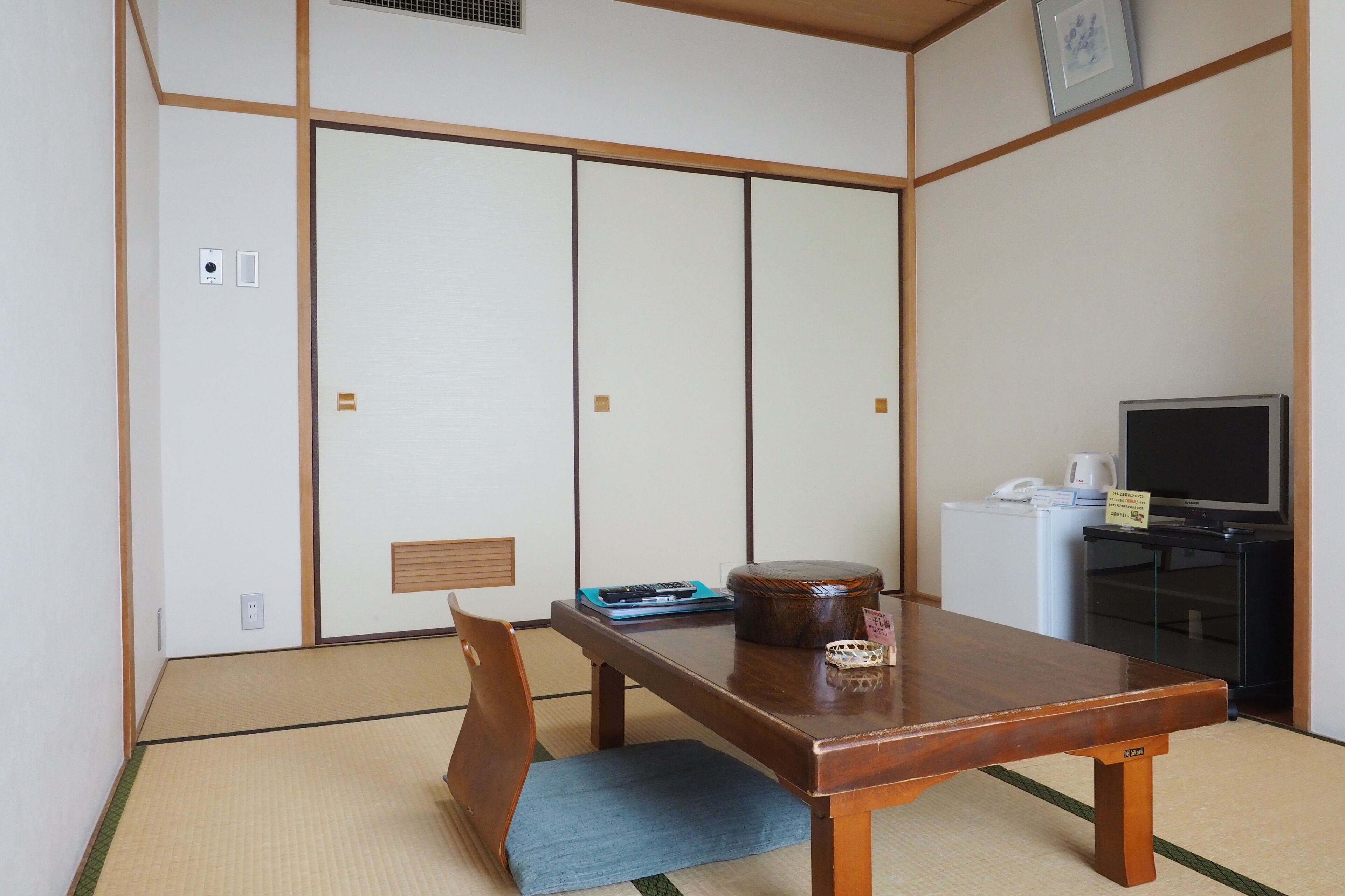 A traditional Japanese room with tatami flooring, a low wooden table, a blue cushion, a television, and a small refrigerator.