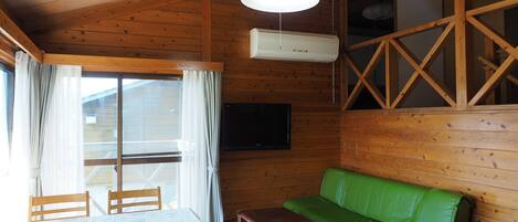A wooden cabin interior with a dining table, chairs, a green sofa, a small table with a book, and a mounted TV.