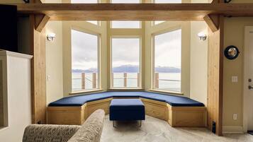 Ein Zimmer mit integriertem Sitzbereich, großen Fenstern und Blick auf ein Gewässer mit Bergen im Hintergrund.