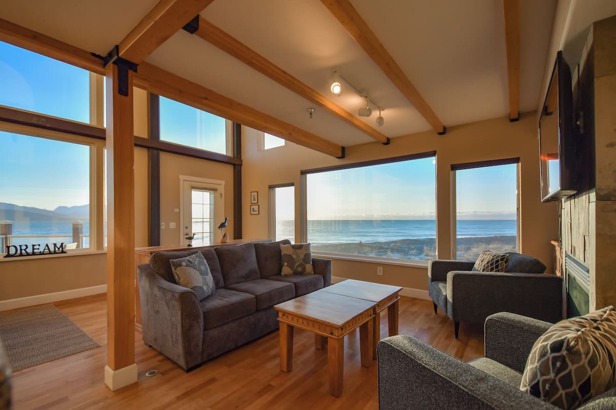 A spacious living room with a large wooden ceiling, a sofa set, a coffee table, and large windows offering ocean views.