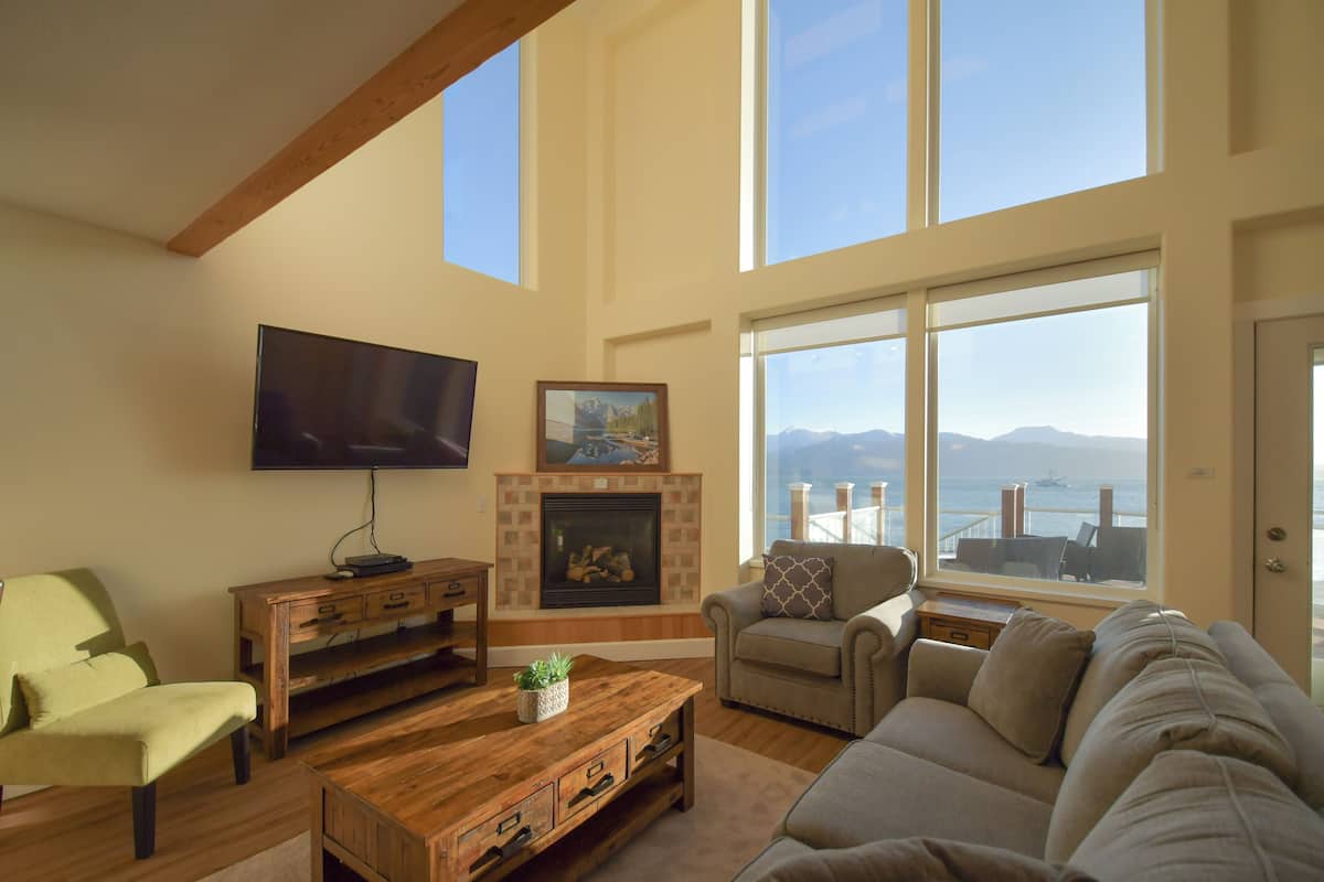 A spacious living room with a fireplace, a television, and a large window offering a view of the sea.