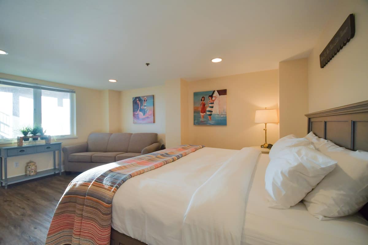 A hotel room with a large bed, a sofa, a desk with a lamp, and artwork on the walls.