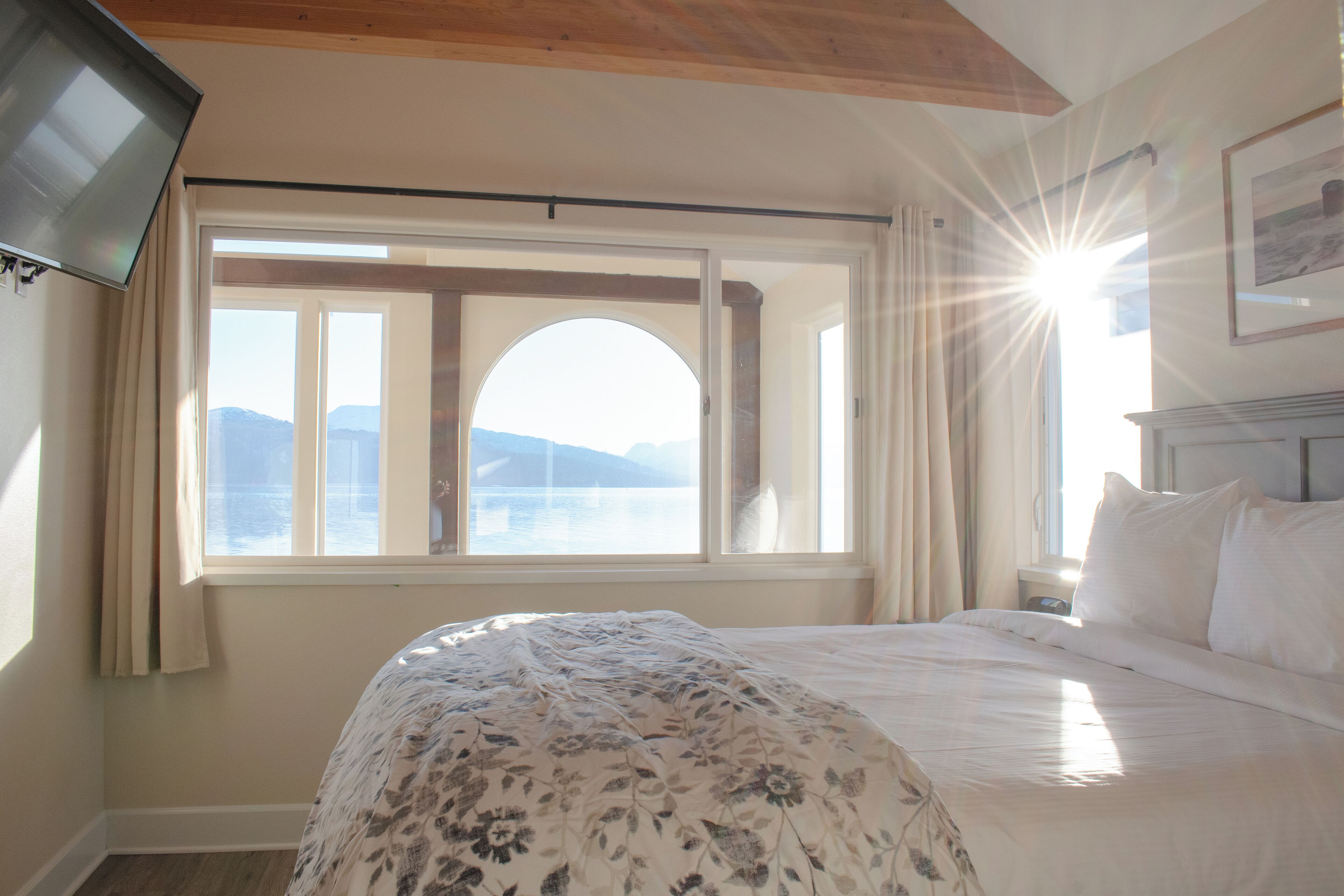 A bedroom with a bed, a window with curtains, a flat-screen TV mounted on the wall, and a framed picture on the wall.