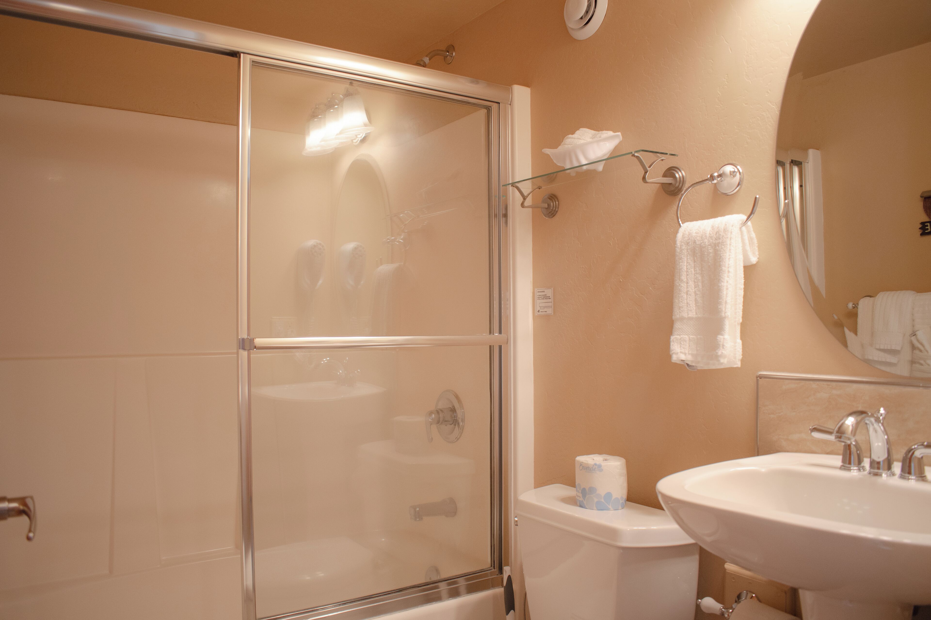 A bathroom with a glass-enclosed shower, a toilet with a paper towel holder, and a sink with a mirror above it.