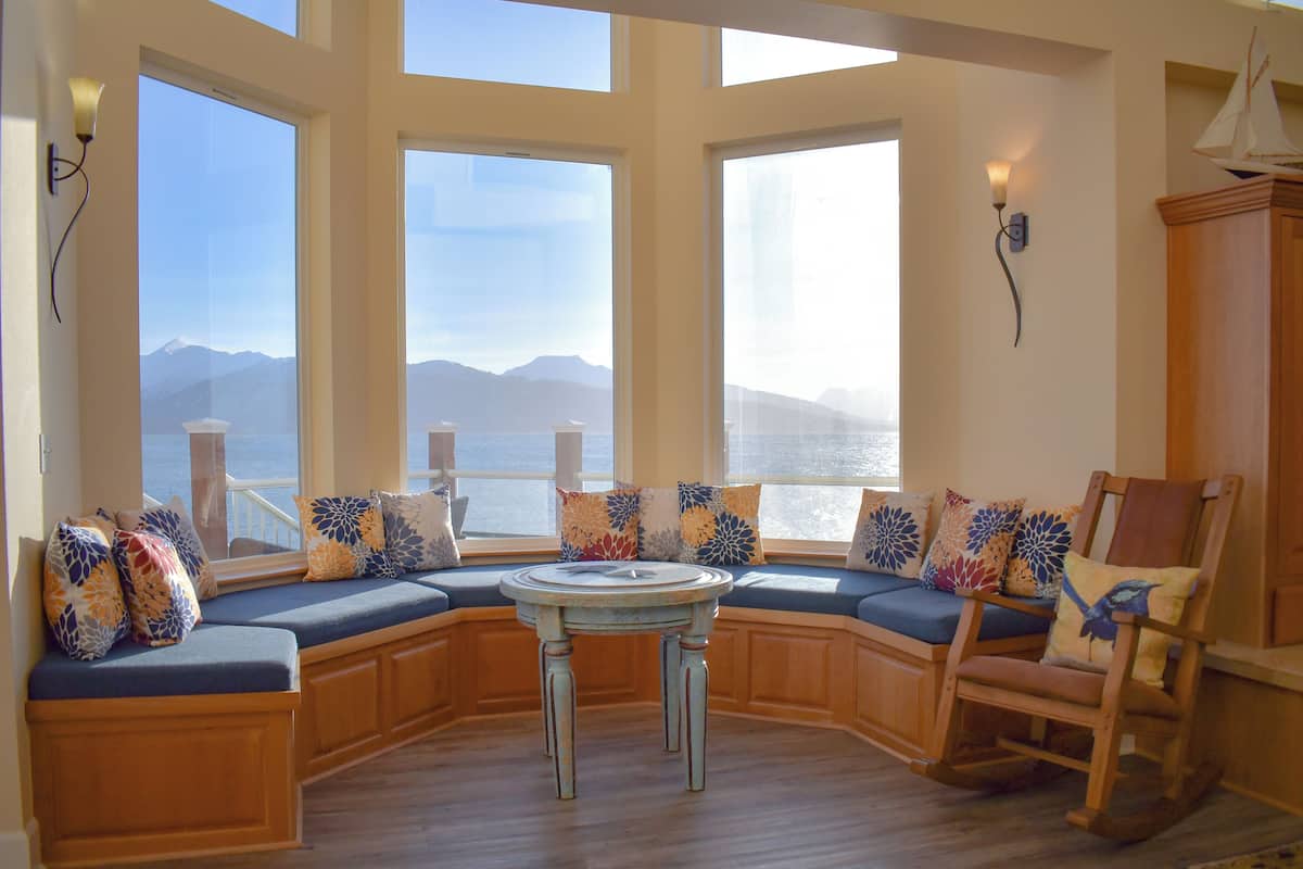 A bay window seating area with a view of the sea and mountains, wooden furniture, and decorative pillows.
