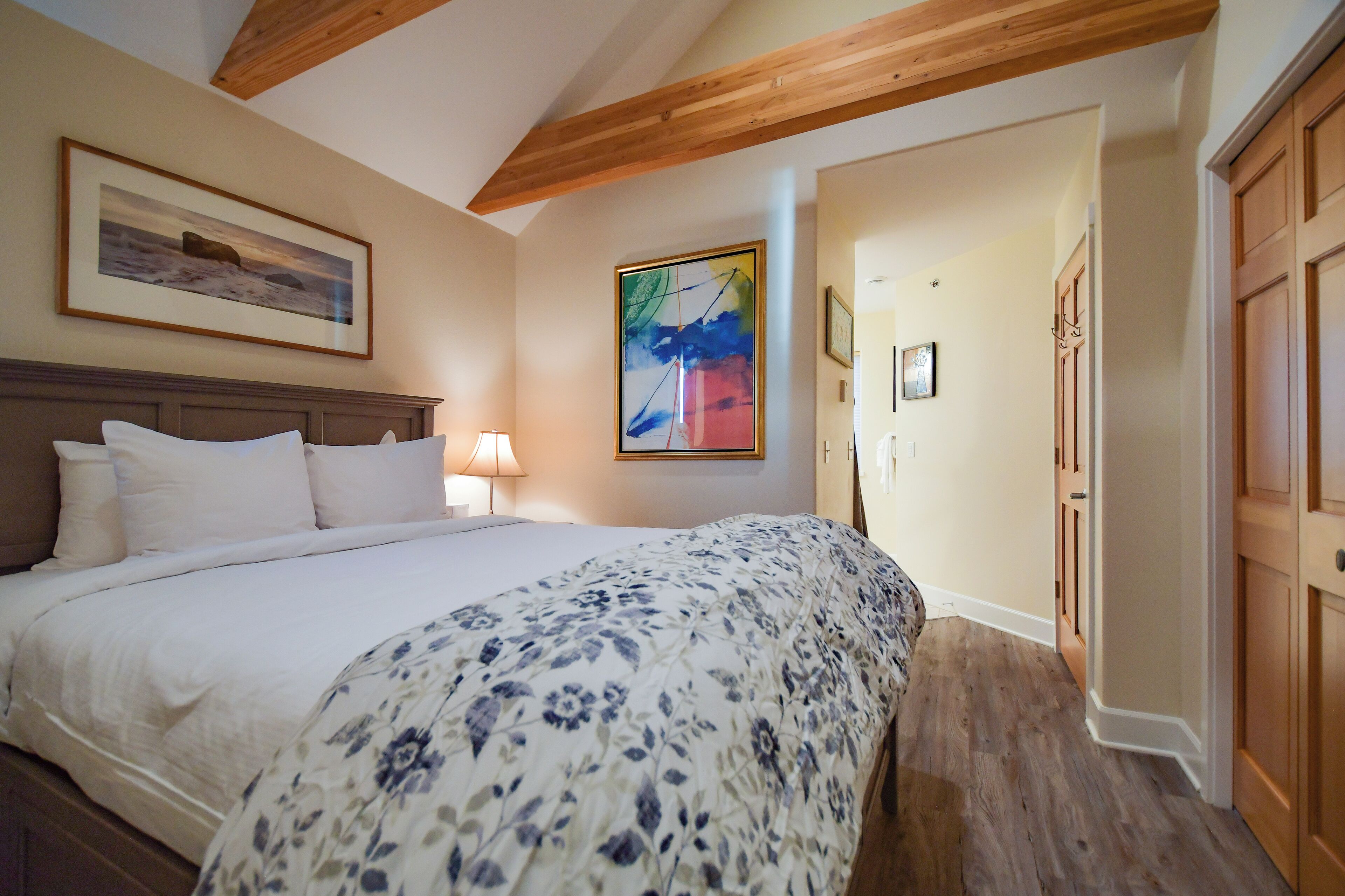 A bedroom with a large bed, wooden ceiling beams, a painting on the wall, and a door to the right.