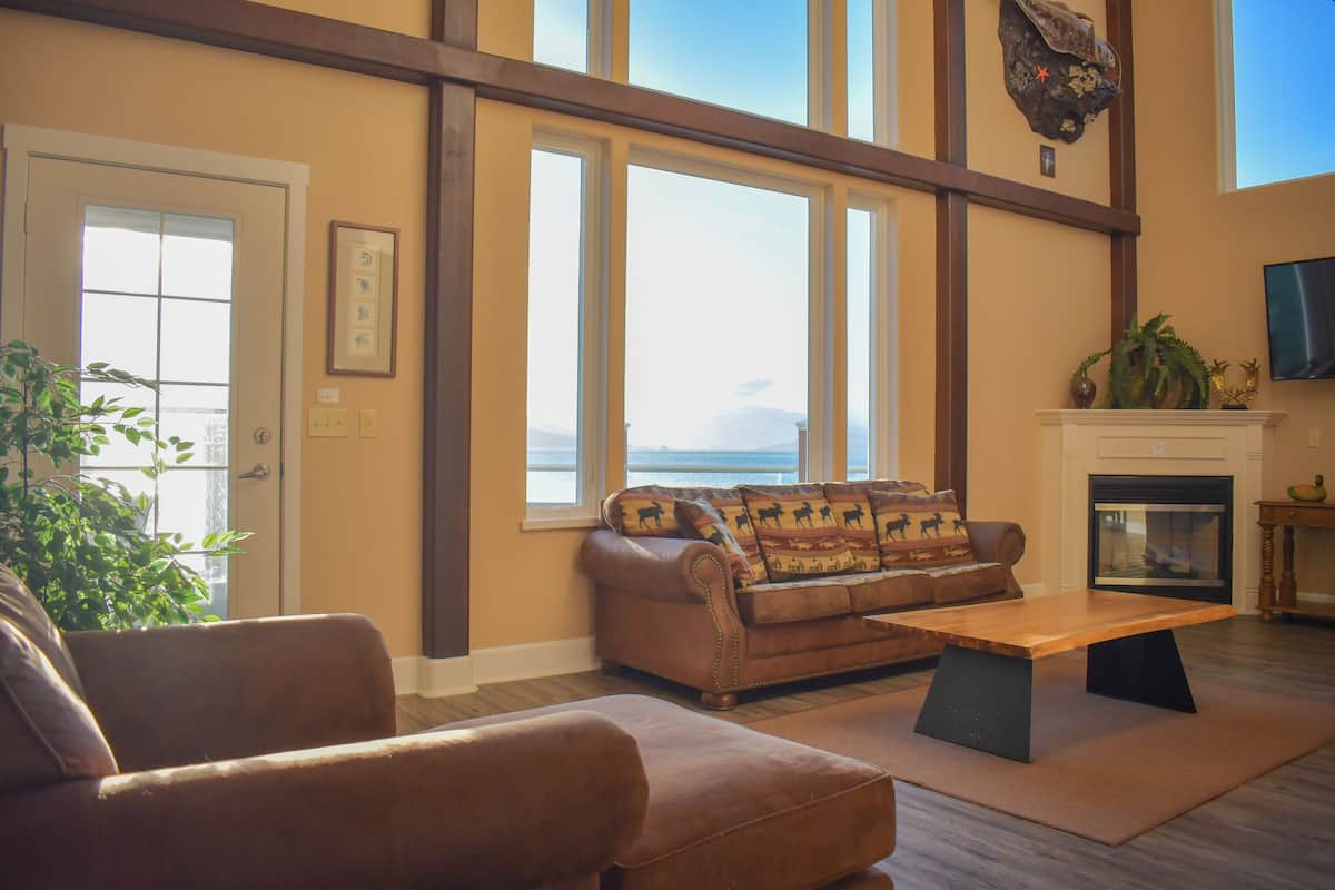 A living room with a sofa, armchair, and coffee table, large windows with a view, a fireplace, and a mounted television.