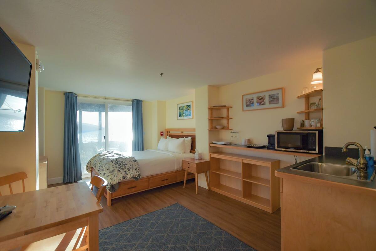 A compact hotel room with a bed, a small desk, a kitchenette, and a large window with curtains.