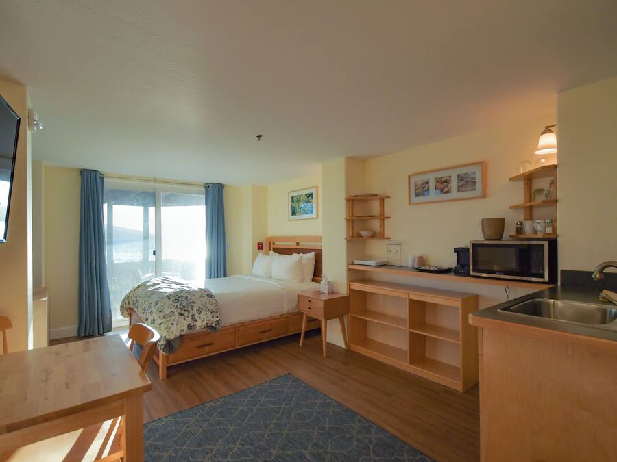 A compact hotel room with a bed, a small desk, a kitchenette, and a large window with curtains.