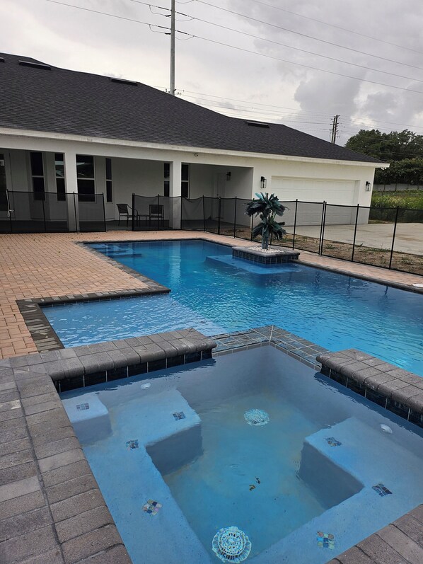 A swimming pool with a hot tub, surrounded by a fence and a building with a white exterior and a dark roof.