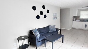 A modern living room with a sofa and a coffee table, a kitchen with white cabinets, and a small round table.