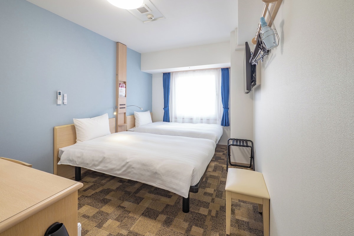 A hotel room with two beds, a desk, a chair, a window with curtains, and a wall-mounted TV.