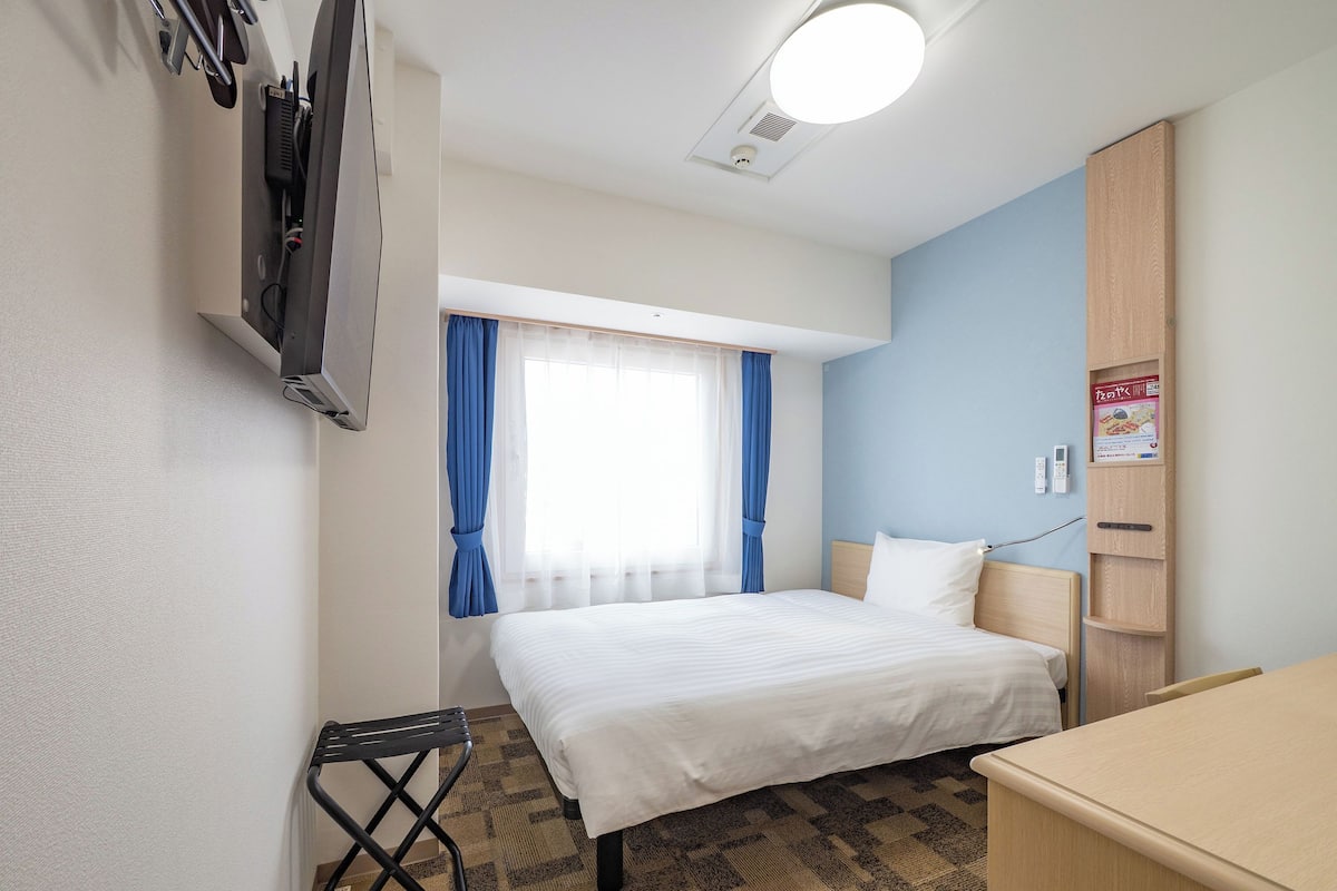 A hotel room with a bed, a desk, a chair, a TV, a window with curtains, and a wall-mounted shelf.