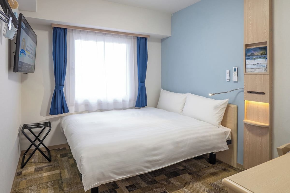 A hotel room with a large bed, a small desk, a window with curtains, a wall-mounted TV, and a bedside table with a lamp.