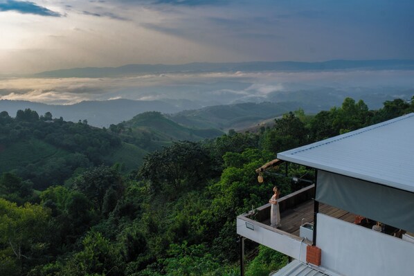 A high-rise building with a balcony overlooking a mountainous landscape with dense greenery and a view of distant clouds.