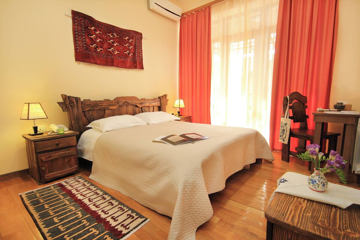 A bedroom with a bed, two bedside tables, a nightstand, a wooden chair, a small table with a vase, and a rug.