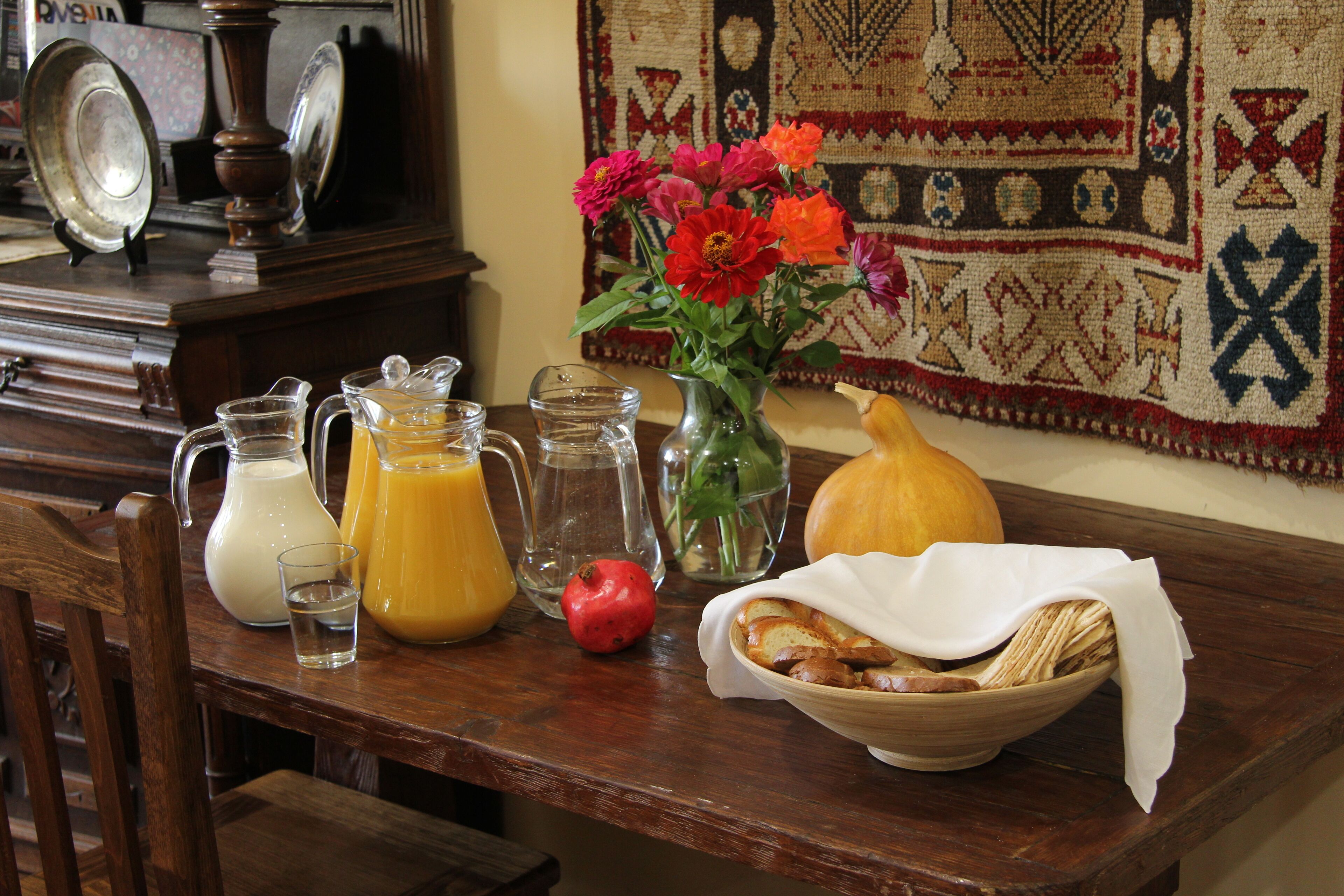 A wooden table with a pitcher of milk, a jug of orange juice, a glass of water, a vase with flowers, a pumpkin, and a bowl of bread.