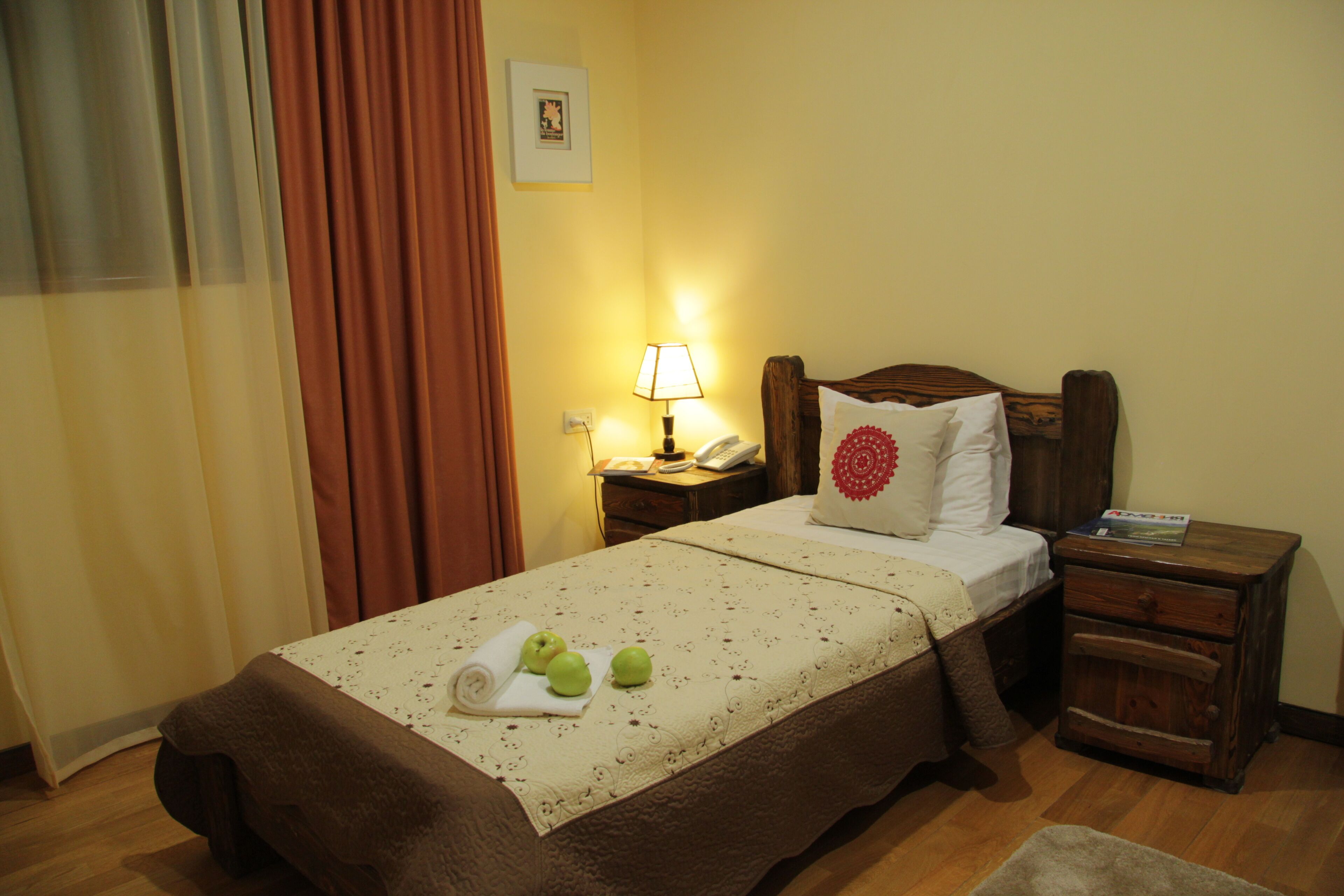 A neatly made bed with a brown bedspread and a decorative pillow, a bedside table with a lamp, and a wooden nightstand with a book.