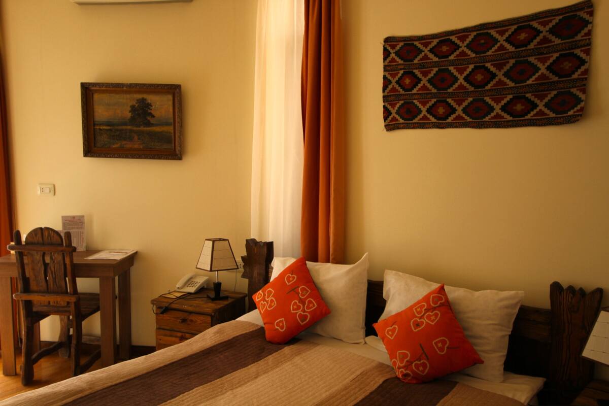 A hotel room with a bed, a desk with a telephone, a chair, and a wall-mounted painting.