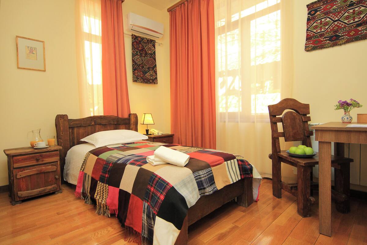 A bedroom with a wooden bed, a nightstand with a lamp, a chair, and a table with a vase of flowers.