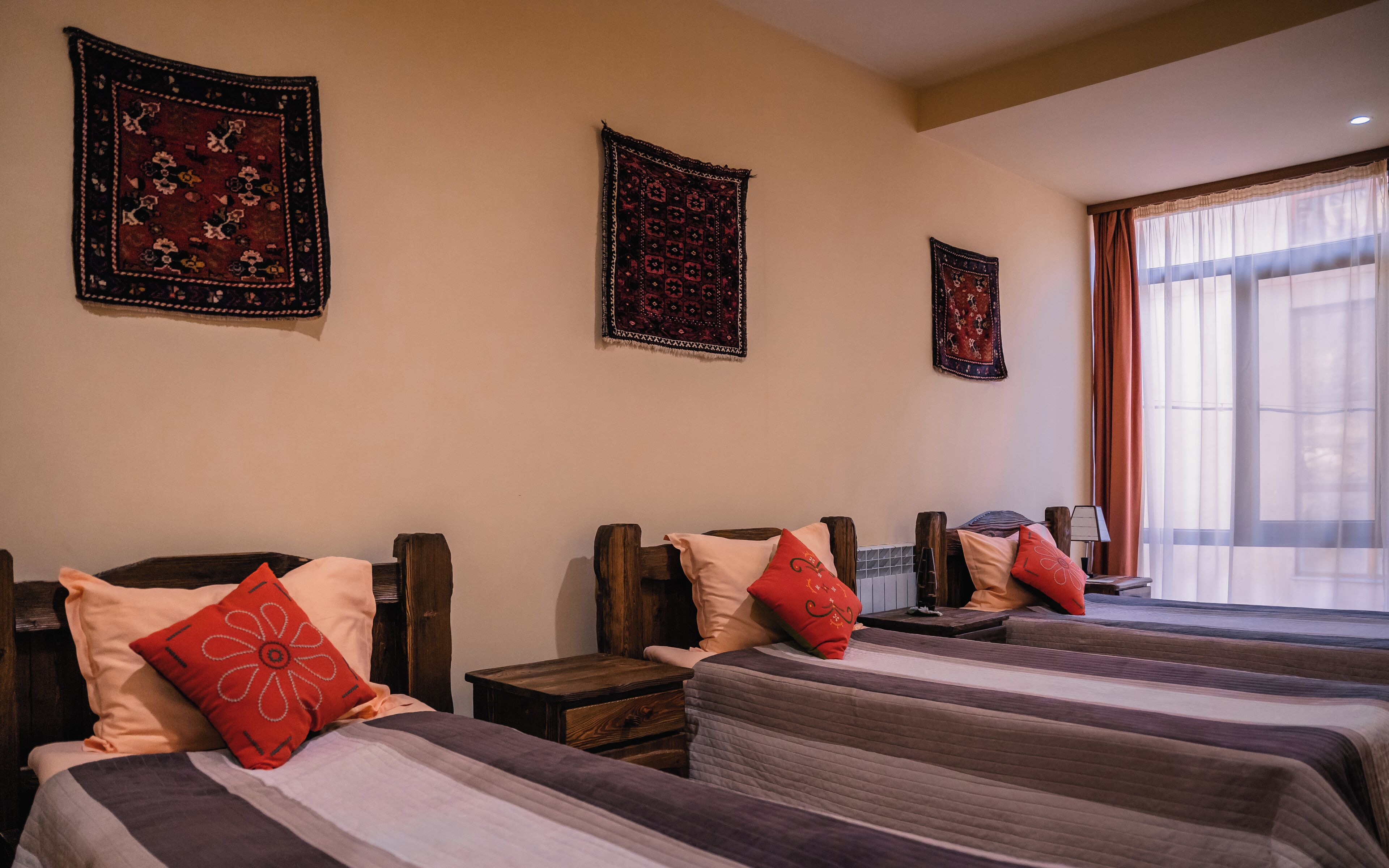 Une chambre d’hôtel avec trois lits, chacun doté d’une tête de lit en bois et d’une couvre-lit à rayures. On y trouve des coussins décoratifs et une petite table de chevet en bois placée entre les lits. La chambre dispose d’une grande fenêtre avec des rideaux légers et offre une vue sur l’extérieur.