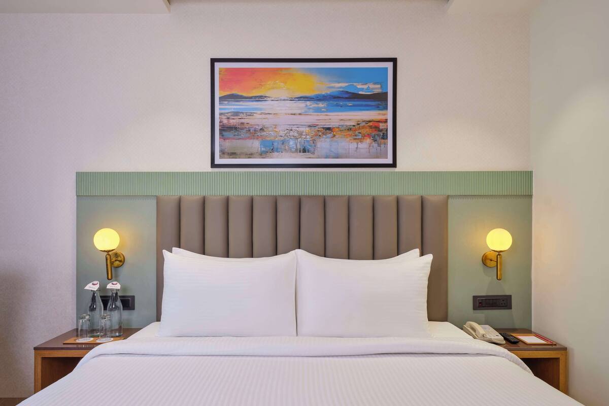 A hotel room with a large bed, two bedside tables with lamps, a framed painting above the bed, and a green headboard.