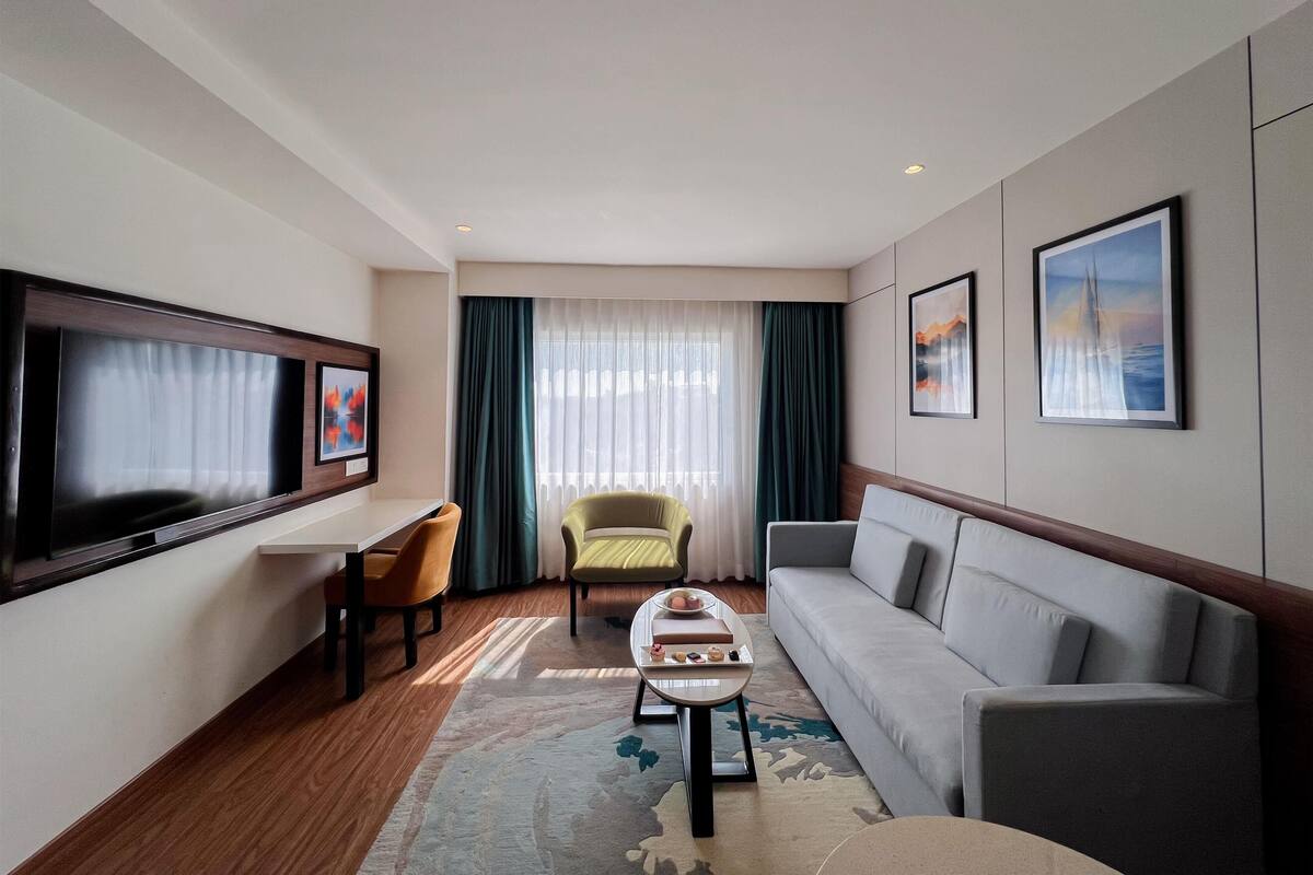 A modern hotel room with a flat-screen TV, a sofa, a chair, a small table, and a large window with curtains.