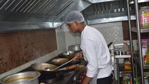 A chef cooking in a stainless steel kitchen with a large wok on a stove, a pot on the counter, and various kitchen utensils and ingredients. - Hotel Yumi Ragi (Papumpare)