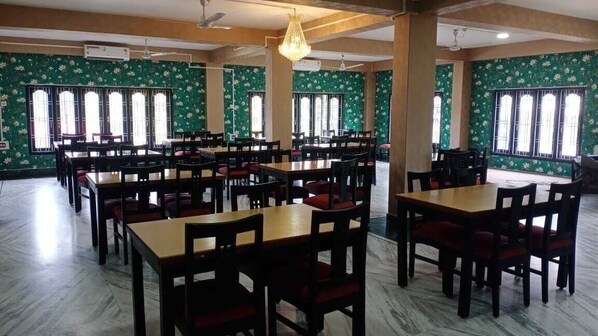 Un'ampia sala da pranzo con diversi tavoli e sedie, grandi finestre con grate decorate e un lampadario.