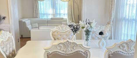 Un coin repas avec une table et des chaises blanches, un lustre et une vue sur un salon comprenant un canapé et des rideaux.