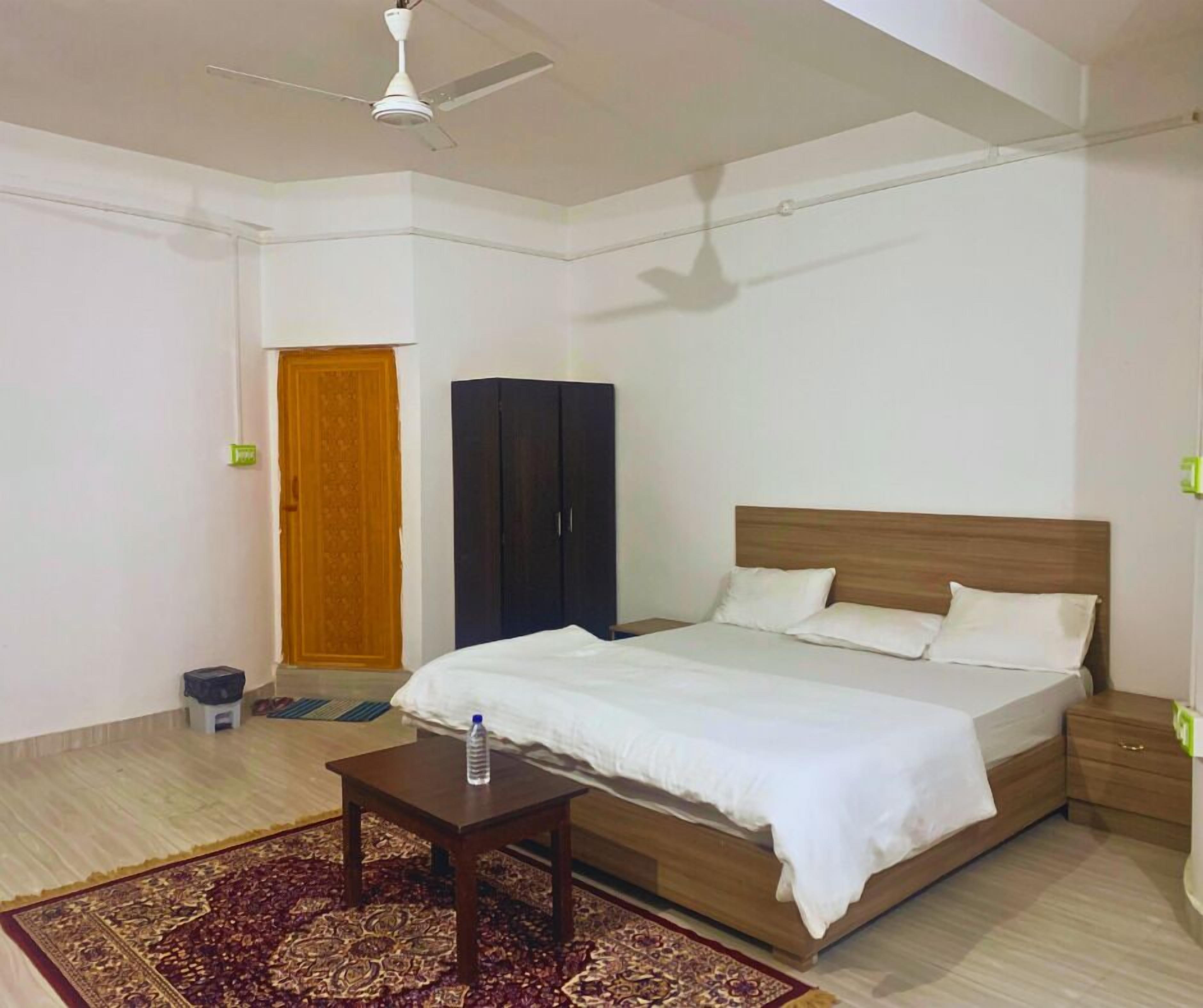 A bedroom with a bed, a wooden wardrobe, a small table with a water bottle, a ceiling fan, and a patterned rug.