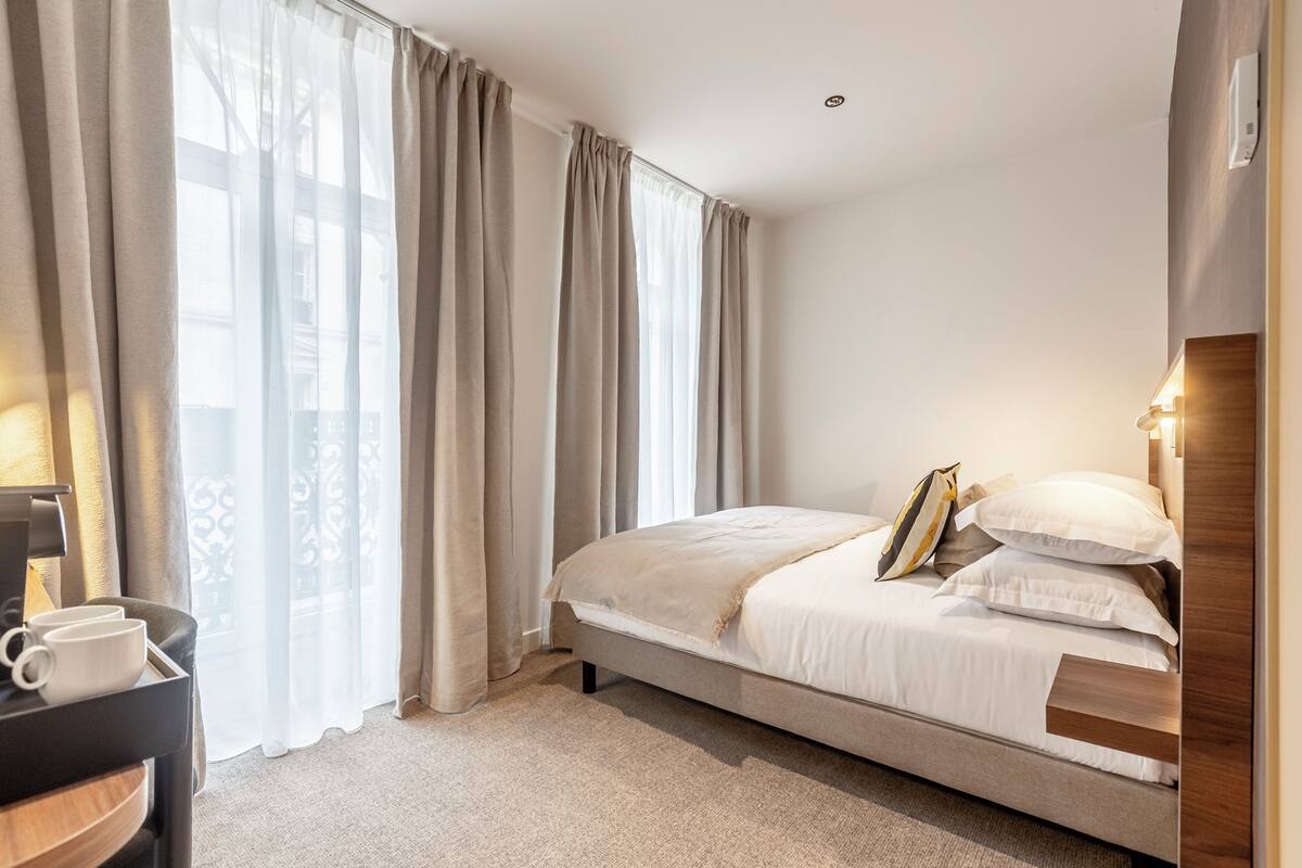 Ein Schlafzimmer mit einem Bett, Vorhängen, einem Nachttisch mit Lampe und einer Kaffeemaschine.