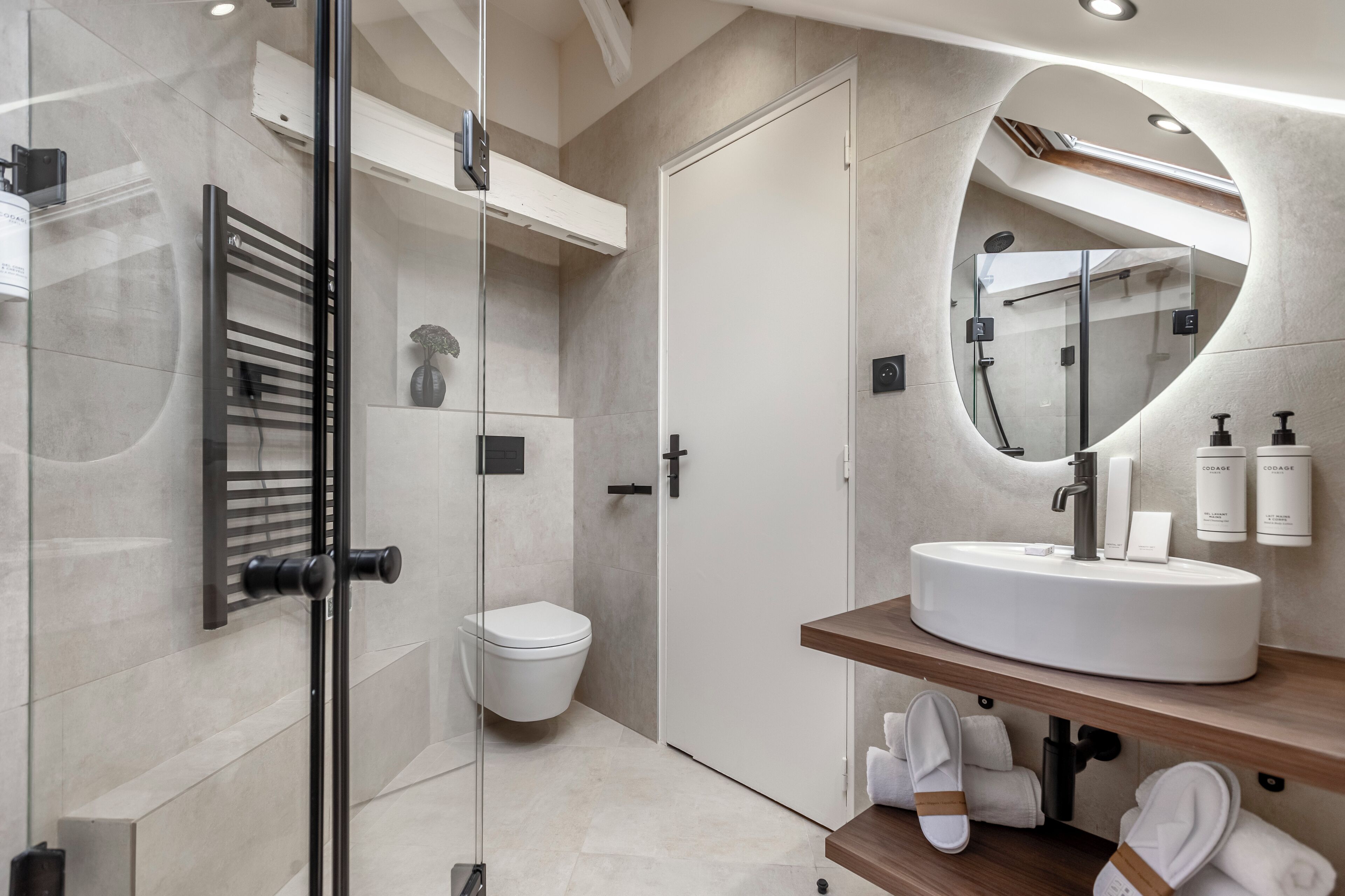 A modern bathroom with a glass shower enclosure, a white toilet, a round mirror, and a wooden vanity with a sink.