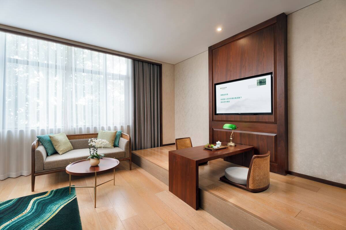Une chambre d’hôtel moderne équipée d’un canapé, d’une table à manger, d’un téléviseur fixé au mur et d’une grande fenêtre avec des rideaux.